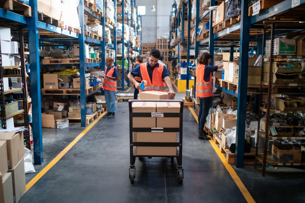 Worker packing cardboard box with tape at on trolley at distribution warehouse